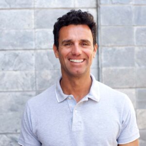 Friendly man smiling outdoors in casual attire against a grey brick wall.
