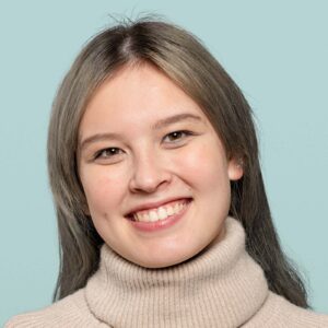 Bright smiling woman with clear skin, long brown hair, wearing a cozy beige turtleneck sweater, portrait, cheerful expression, friendly face, natural makeup, light blue background.