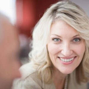 Friendly blonde woman smiling, engaging in conversation, customer service, professional communication, modern office setting, business collaboration, positive attitude, professional demeanor, social interaction, corporate environment.