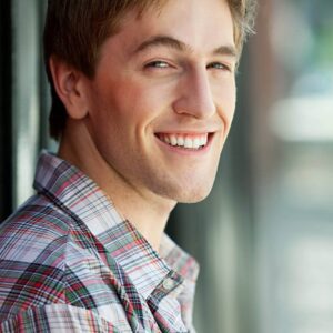 Smiling young man in casual plaid shirt looking out of window, natural light, happy expression, modern office setting, friendly professional.