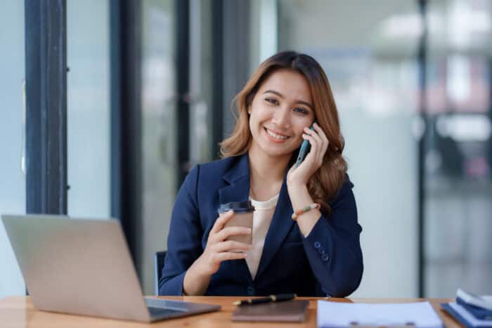 A businesswoman smiling while talking on a mobile phone at her desk with a laptop and coffee cup, representing professional communication and modern office work.
