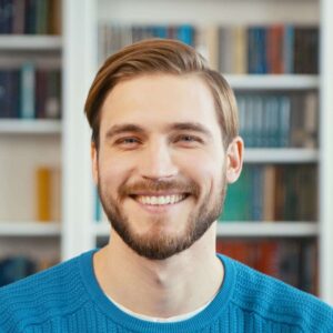 Smiling young man in blue sweater in a library with shelves of books in the background.