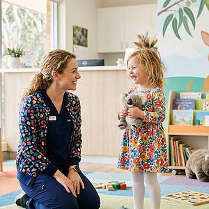 A caregiver and young girl smiling and playing together in a colorful childcare center with toys, books, and animal-themed wall decor, promoting a friendly and nurturing environment.